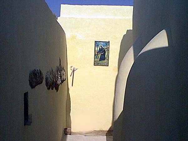 Outdoor alleyway between two pale yellow walls under a blue sky, with a small painted religious icon mounted on the wall