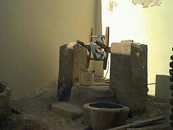 Interior scene of an old stone well or water mechanism with a wooden frame, sandy floor, warm brown and beige tones, rustic mood