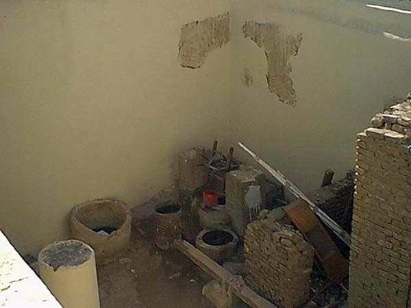 Rooftop courtyard corner with plain plastered walls, patched stains, and large ceramic vessels and building materials scattered about