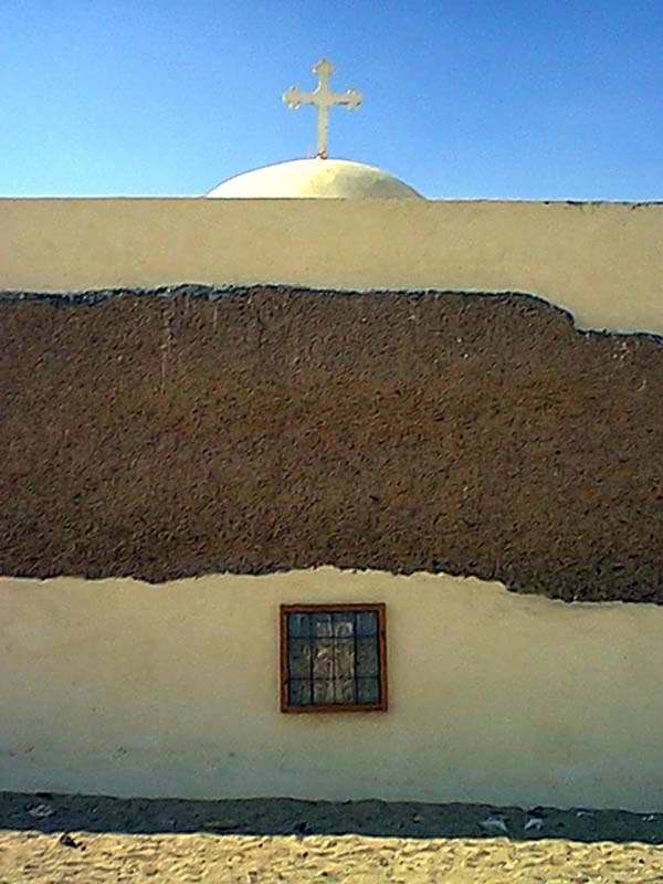 Domed building exterior with a cross on top, textured brown and cream wall sections, and a small barred window under bright sky