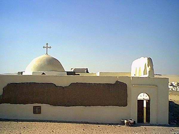Small domed building with a cross on top, whitewashed walls with a section of exposed mud brick, standing under a clear blue desert sky